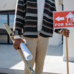 A real estate agent stands outside with a 'for sale' sign and rolled blueprint, suggesting a property for sale.